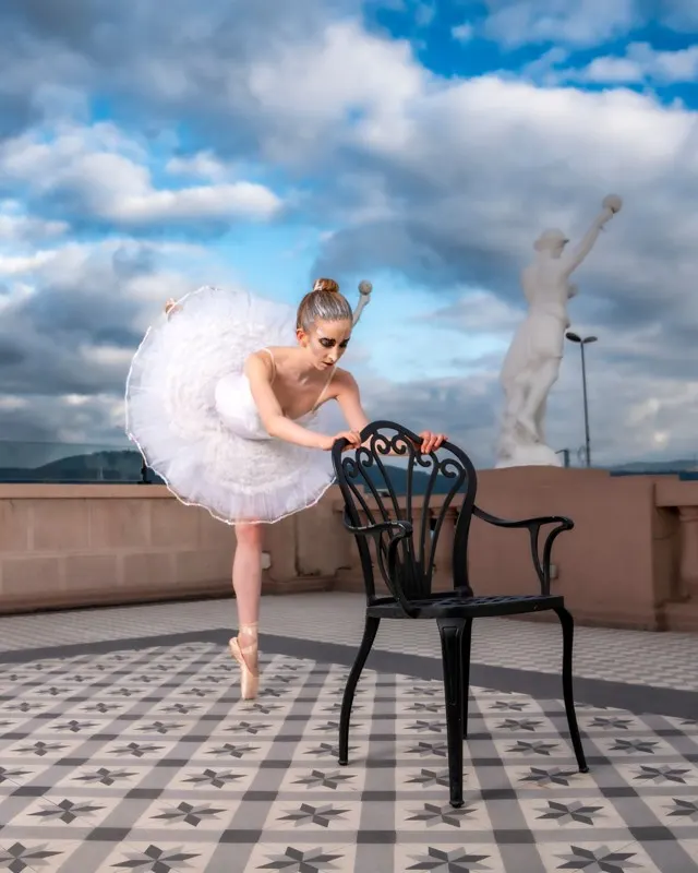 Bailarina con tutú apoyada en una silla, en una terraza bajo nubes.